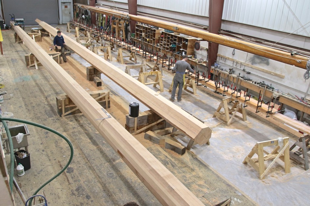 On the right side of the photo Kaz Buozys is checking clamps on the recent lamination for the main boom on the far side of the shop floor. David Short (blue shirt) is seen here wielding his template for shaping the foremast (on the right).  The main mast (left) is already 8 sided in this image.   Soon, the main and foremast will be completed and ready for coating before hardware is installed.