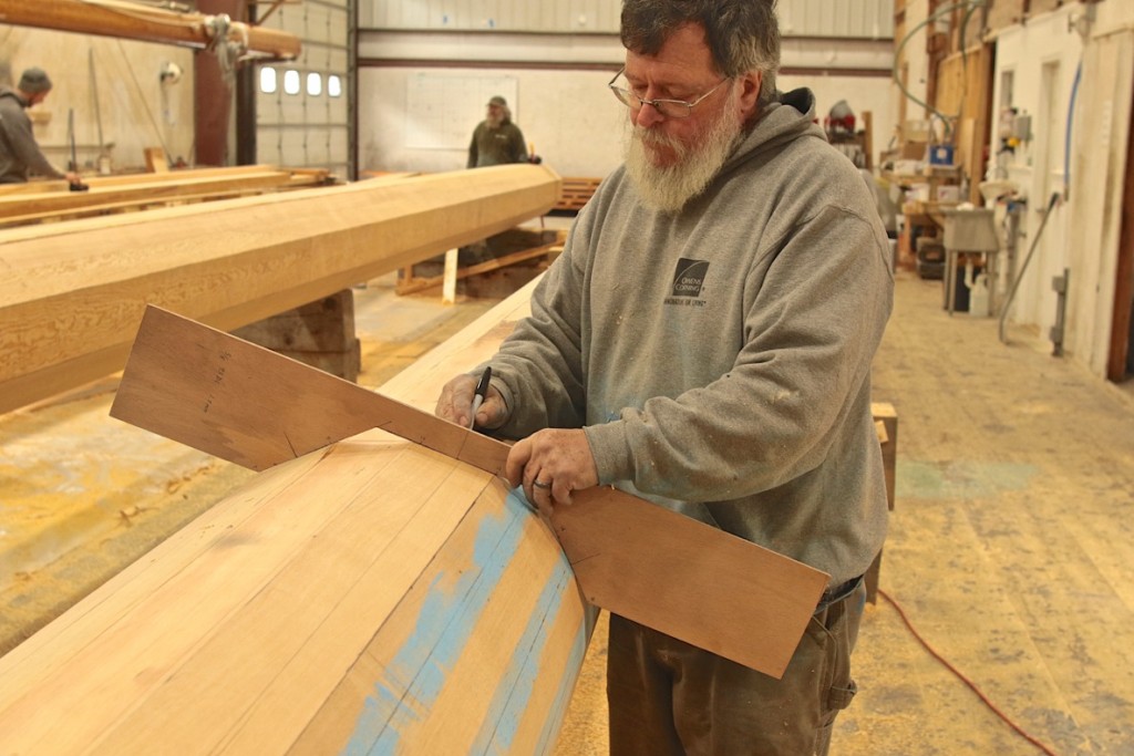 David Short monitoring the shaping process.  You can see in this image the progress of bringing the Main Mast from 8-sides to 16-sides.  This process begins with a chainsaw to rough-cut the mast from a square laminated block into an 8-sided spar.  From that point, the crew uses power planes to bring it as closely to round as possible before sandpaper finishes the job.  In this image, sandpaper is not far off.